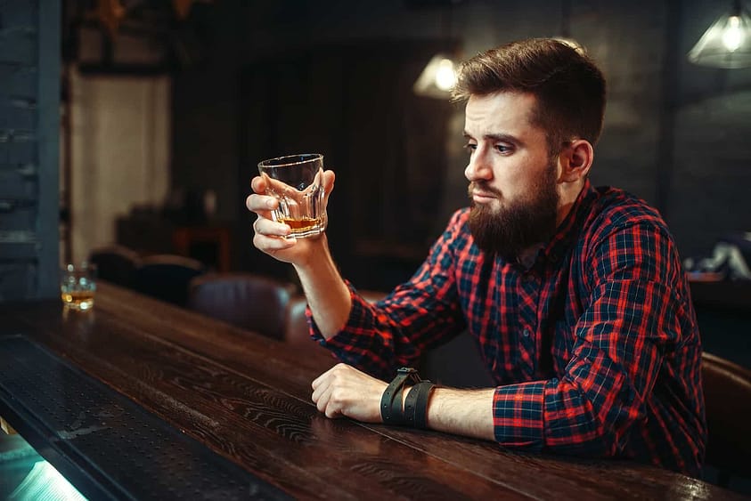 A young man sits at a bar with a drink in his hand, contemplating if he's an alcoholic.