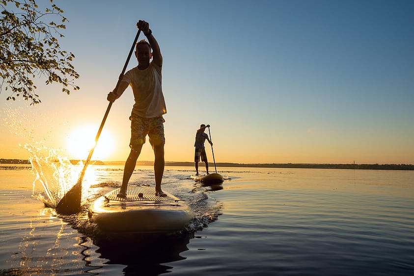 Two men enjoy the benefits of recreational therapy as they paddle board at sunrise.