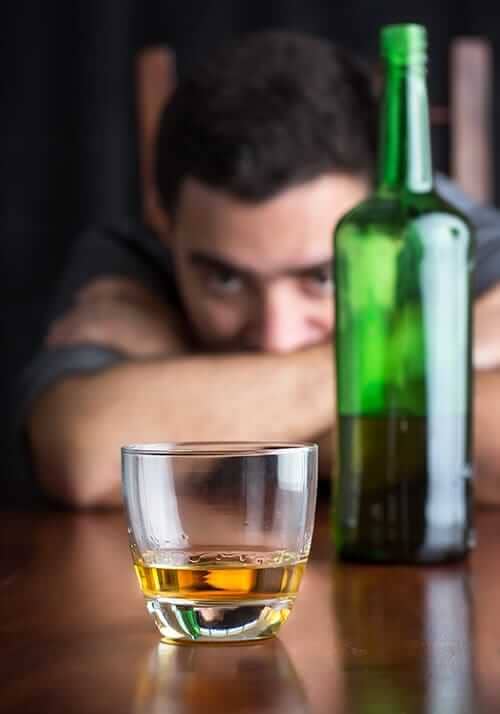 A man rests his head on his arms and stares at a glass of alcohol while he thinks about quitting drinking