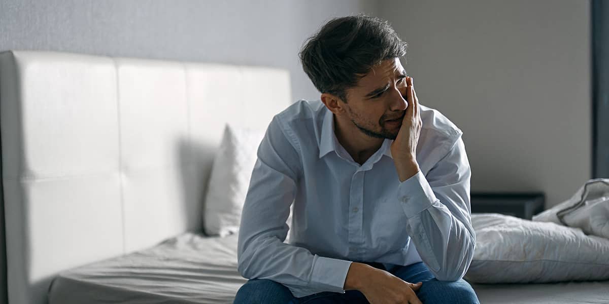 man sitting on bed rubbing face irritated by Alcohol Dependency