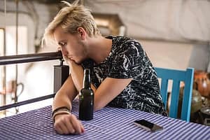 Sad young man at table suffering from alcohol poisoning is disparate to quit.