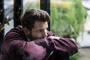 Depressed man looking out rainy window and wondering if he has delirium tremens