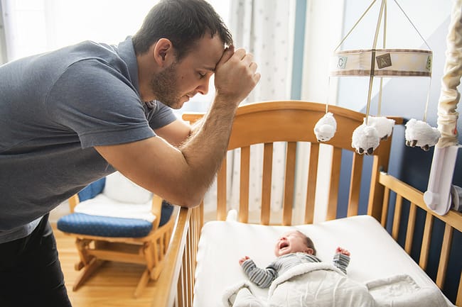 man leaning against his baby's crib suffering from symptoms of postpartum depression for men
