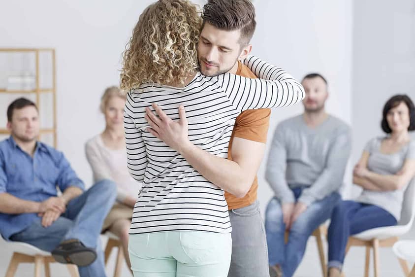 A young man and woman comfort one another during group therapy by hugging.